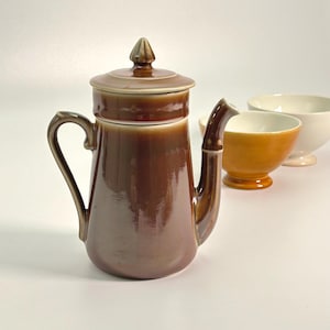 Puede incluir: Una cafetera de cerámica marrón con tapa y asa curva. La jarra tiene un acabado brillante y está acompañada de un cuenco naranja pequeño y un cuenco blanco. Los artículos están dispuestos sobre una superficie blanca.