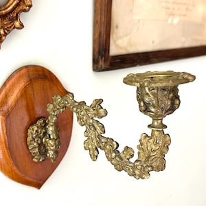 May include: A wall-mounted candle holder featuring a polished brass arm and cup decorated with detailed oak leaf and acorn motifs. The brass fixture is securely attached to a smooth, shield-shaped wooden plaque with a warm, light brown finish.