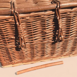 May include: A close-up of a brown wicker picnic basket with a wooden handle and metal clasps. The basket is intricately woven with a natural, textured appearance. A wooden dowel lies in front of the basket.