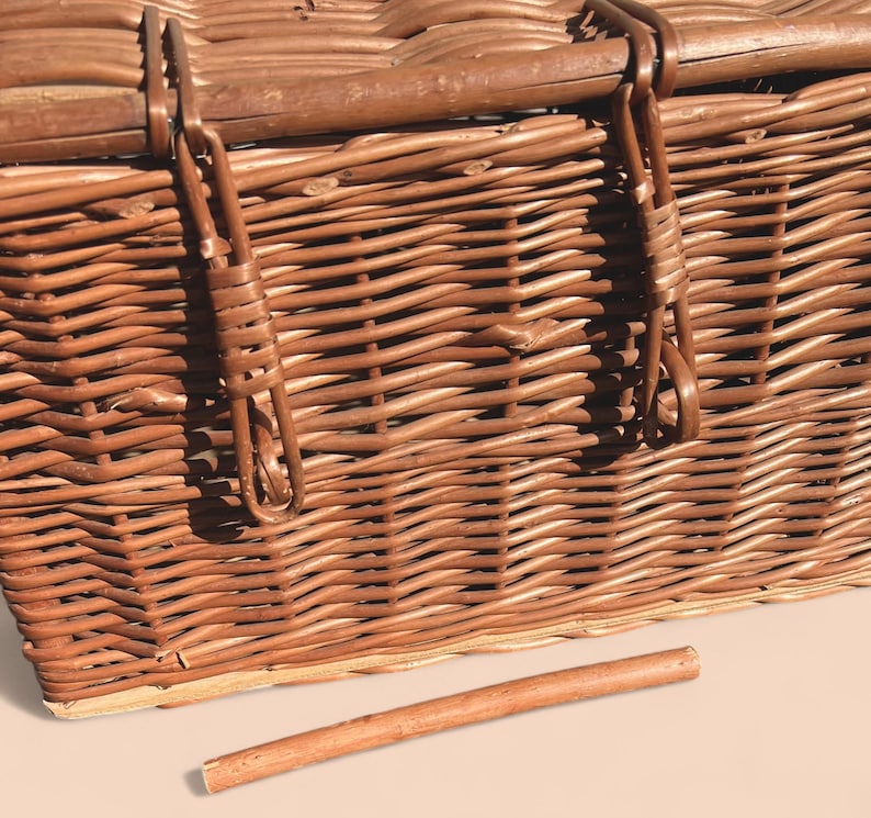 May include: A close-up of a brown wicker picnic basket with a wooden handle and metal clasps. The basket is intricately woven with a natural, textured appearance. A wooden dowel lies in front of the basket.
