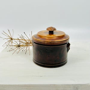 May include: A brown ceramic lidded jar with a decorative knob on top. The jar has a dark brown base and a lighter brown lid. The jar has two small handles on the sides. The jar is sitting on a white surface.