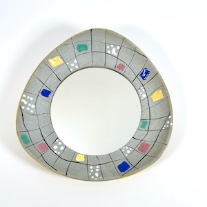 May include: A decorative wall mirror with an abstract design. The grey frame is irregularly shaped and features a grid pattern with colourful square accents in blue, yellow, pink, and green. White polka dots add detail to the design.