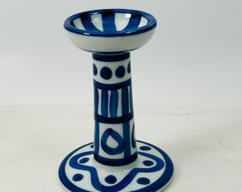 May include: A ceramic candle holder with a white base and a blue and white design. The candle holder has a round base, a cylindrical stem, and a small bowl-shaped top. The design includes stripes, dots, and teardrop shapes.