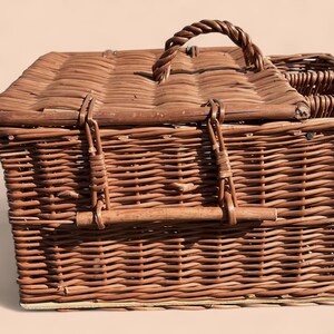 May include: A brown wicker picnic basket with a woven handle and a hinged lid. The basket has metal clasps and a woven design. The basket is made of natural materials and is designed for carrying food and drinks.
