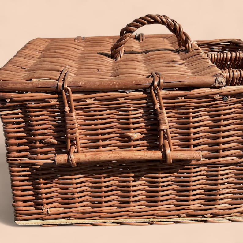 May include: A brown wicker picnic basket with a woven handle and a hinged lid. The basket has metal clasps and a woven design. The basket is made of natural materials and is designed for carrying food and drinks.