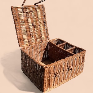 May include: A rectangular wicker picnic basket with a hinged lid, open to reveal interior compartments. The basket is woven with brown and tan reeds, featuring a handle on the lid and side. The interior includes dividers for organizing items.