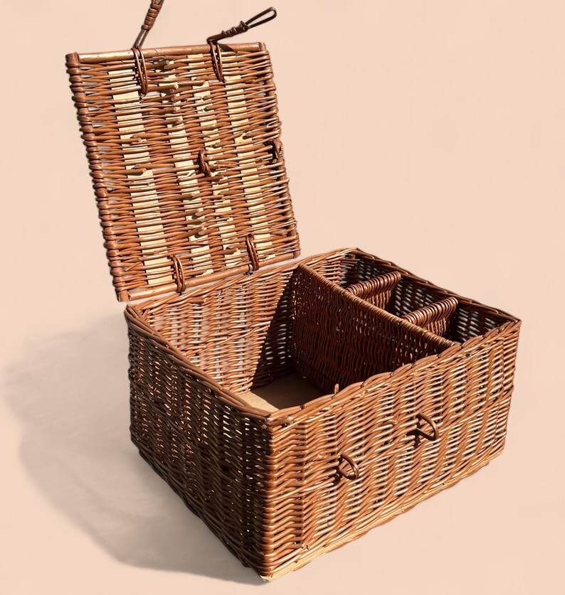 May include: A rectangular wicker picnic basket with a hinged lid, open to reveal interior compartments. The basket is woven with brown and tan reeds, featuring a handle on the lid and side. The interior includes dividers for organizing items.