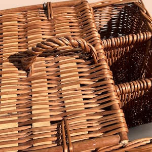 May include: Close-up of a brown wicker basket with a woven handle. The basket has a rectangular shape and is made of interwoven strips of wood. The handle is made of twisted wicker and is attached to the top of the basket.