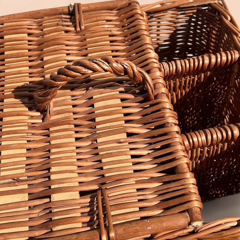 May include: Close-up of a brown wicker basket with a woven handle. The basket has a rectangular shape and is made of interwoven strips of wood. The handle is made of twisted wicker and is attached to the top of the basket.