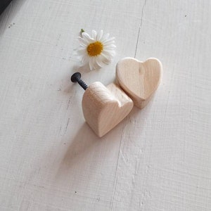 Könnte beinhalten: Zwei unfertige, herzförmige Schubladenknöpfe aus Holz mit einer schwarzen Schraube auf einer weißen Oberfläche.