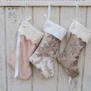 May include: Three Christmas stockings hanging on a white wooden wall. The stockings are made of fabric and have lace trim. One stocking is pink, and the other two are a beige color with a floral pattern.