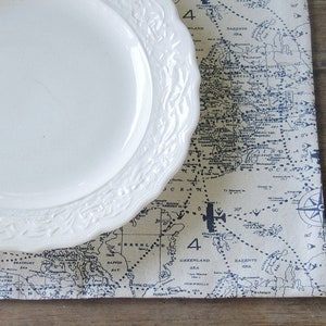 May include: A white, scalloped-edge plate rests on a map-themed placemat. The placemat features a vintage map design in blue and beige, with nautical elements. The plate's rim has a detailed, embossed pattern. The background is a wooden surface.