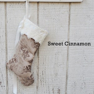 May include: A brown and beige Christmas stocking with a white lace trim and a white ribbon. The stocking is hanging on a white wooden wall with the text "Sweet Cinnamon" written below it.