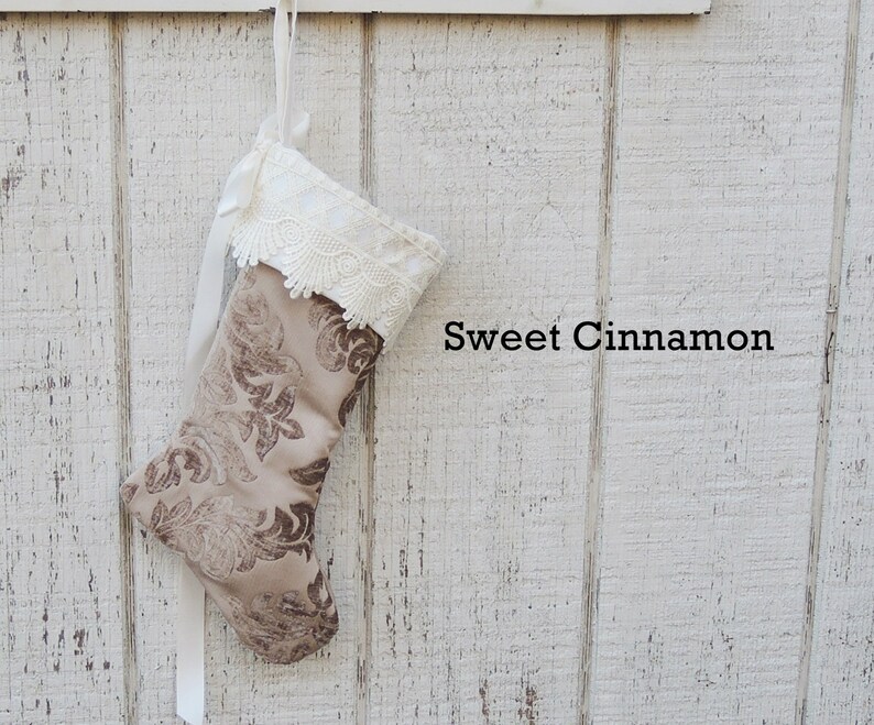 May include: A brown and beige Christmas stocking with a white lace trim and a white ribbon. The stocking is hanging on a white wooden wall with the text "Sweet Cinnamon" written below it.
