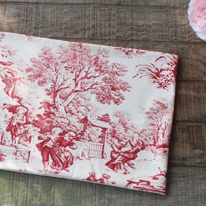 May include: A folded piece of fabric with a red and white toile pattern. The design depicts figures in a pastoral scene with trees and buildings. The fabric is resting on a wooden surface, with pink flowers in the background.