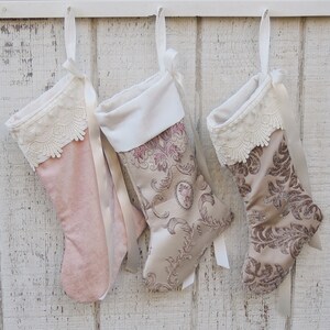 May include: Three Christmas stockings hanging on a white wooden wall. The stockings are made of velvet and have a lace trim. The stockings are decorated with a floral pattern.