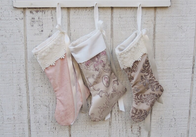 May include: Three Christmas stockings hanging on a white wooden wall. The stockings are made of velvet and have a lace trim. The stockings are decorated with a floral pattern.