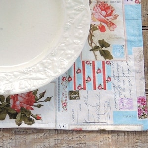 May include: A white porcelain plate with a floral design sits on top of a blue and white patterned placemat with a floral design. The placemat has a handwritten message and a stamp.