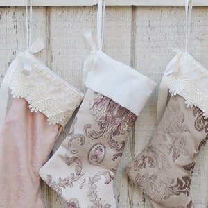 May include: Three Christmas stockings hanging on a white wooden wall. The stockings are made of different fabrics and have lace trim. One stocking is pink, one is beige with a floral pattern, and the other is beige with a paisley pattern.