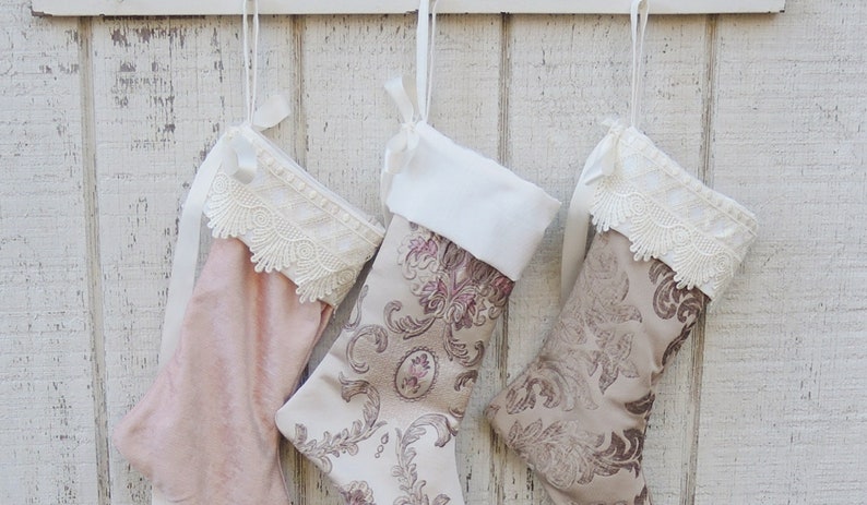 May include: Three Christmas stockings hanging on a white wooden wall. The stockings are made of different fabrics and have lace trim. One stocking is pink, one is beige with a floral pattern, and the other is beige with a paisley pattern.
