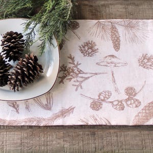 May include: A rectangular fabric table runner featuring a light pink background with a repeating brown botanical print of pinecones, pine needles, and mushrooms. A white square plate holds three brown pinecones, with green pine branches nearby.