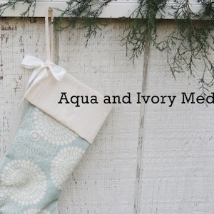 May include: A Christmas stocking with a white cuff and a blue and white floral pattern. The stocking is hanging on a white wooden wall and has a white ribbon tied around the cuff. The text "Aqua and Ivory Medallion" is printed on the stocking.