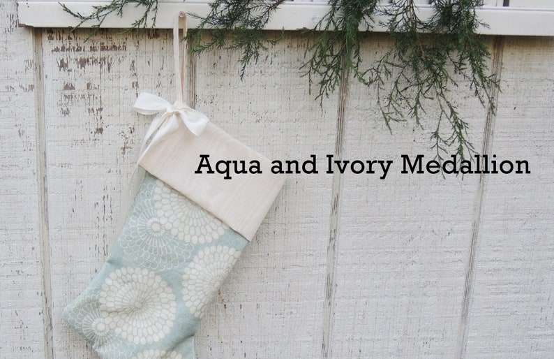 May include: A Christmas stocking with a white cuff and a blue and white floral pattern. The stocking is hanging on a white wooden wall and has a white ribbon tied around the cuff. The text "Aqua and Ivory Medallion" is printed on the stocking.