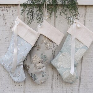 May include: Three blue and white Christmas stockings with white satin ribbon and a cream colored cuff. The stockings are hanging on a white wooden wall.