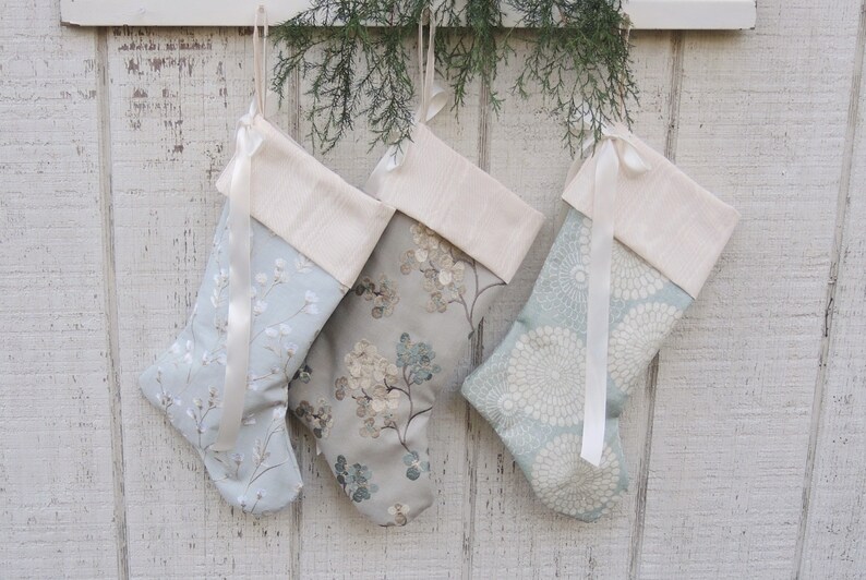 May include: Three blue and white Christmas stockings with white satin ribbon and a cream colored cuff. The stockings are hanging on a white wooden wall.