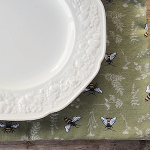 May include: A white ceramic plate with a raised floral design sits on a green placemat with a repeating pattern of bees and foliage.