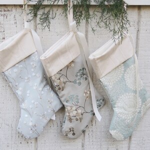 May include: Three Christmas stockings with a white cuff and a blue and white floral pattern. The stockings are hanging on a white wooden wall.