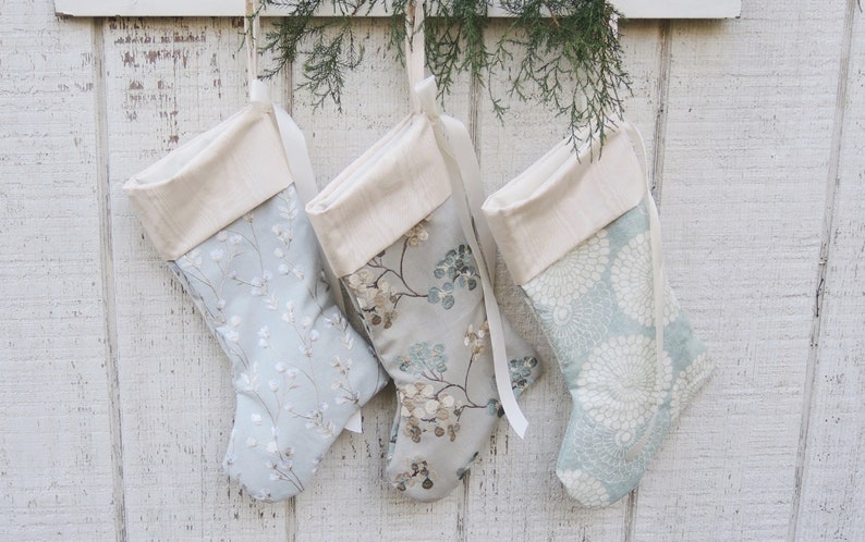 May include: Three Christmas stockings with a white cuff and a blue and white floral pattern. The stockings are hanging on a white wooden wall.