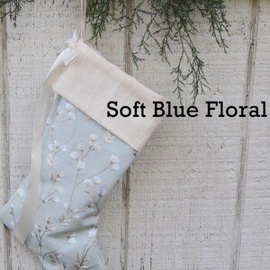 May include: A light blue Christmas stocking with a white floral pattern and a white cuff. The stocking has a white satin ribbon tied around the cuff. The text "Soft Blue Floral" is printed on the stocking.