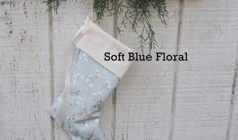 May include: A light blue Christmas stocking with a white floral pattern and a white cuff. The stocking has a white satin ribbon tied around the cuff. The text "Soft Blue Floral" is printed on the stocking.