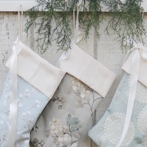 May include: Three white and blue Christmas stockings with white satin ribbon ties hanging on a white wooden wall. The stockings have different patterns, including floral and geometric designs.