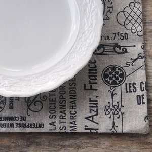 May include: A white ceramic plate with a decorative border sits on a gray and black placemat with French text and designs. The text includes "'Azur, France" and "rix: 7.50".