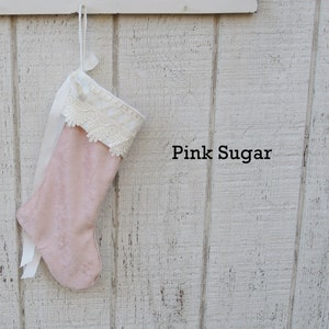 May include: Pink velvet Christmas stocking with white lace trim and a white satin ribbon. The stocking is hanging on a white wooden wall with the text "Pink Sugar" written below it.