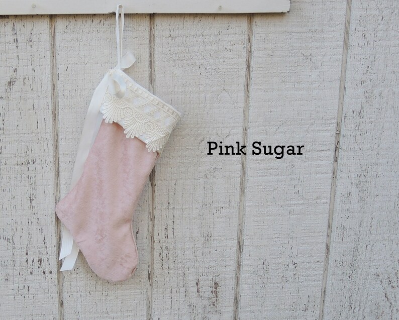 May include: Pink velvet Christmas stocking with white lace trim and a white satin ribbon. The stocking is hanging on a white wooden wall with the text "Pink Sugar" written below it.
