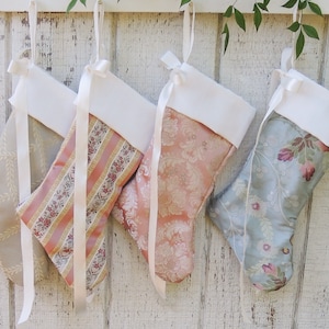 May include: Four Christmas stockings with white faux fur cuffs and satin ribbon ties. The stockings are made of different patterned fabrics in shades of beige, pink, and blue.