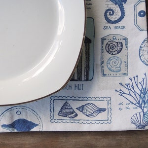 May include: A white ceramic plate with a gold rim sits on a blue and white placemat with a nautical theme. The placemat features images of seashells, seahorses, and a beach hut.