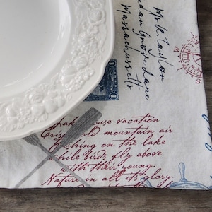 May include: A white ceramic plate with a floral design sits on a white fabric placemat with blue and red text. The text includes the words "vacation", "mountain air", "fishing on the lake", and "Nature in all its glory".