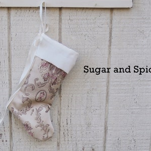 May include: A white and beige Christmas stocking with a floral pattern hangs on a white wooden wall. The stocking has a white cuff and a white ribbon. The text "Sugar and Spice" is written on the wall.