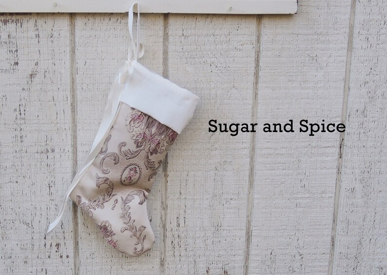 May include: A white and beige Christmas stocking with a floral pattern hangs on a white wooden wall. The stocking has a white cuff and a white ribbon. The text "Sugar and Spice" is written on the wall.