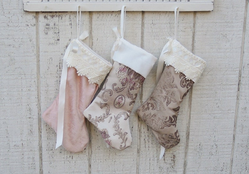 May include: Three Christmas stockings hanging on a white wooden wall. The stockings are made of fabric and have lace trim. One stocking is pink, and the other two are a beige color with a floral pattern.