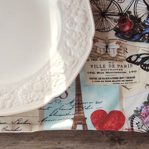 May include: A white porcelain plate with a floral design sits on top of a fabric placemat with a Parisian theme. The placemat features a bicycle, butterflies, and the text "VILLE DE PARIS" with an address. The placemat is decorated with a red heart and a vintage-style Eiffel Tower.
