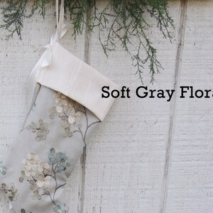 May include: A gray and white Christmas stocking with a floral pattern. The stocking has a white cuff and is hanging on a white wooden wall. The text "Soft Gray Floral" is printed on the wall.
