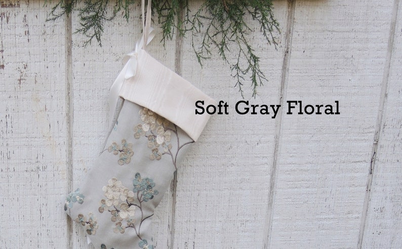 May include: A gray and white Christmas stocking with a floral pattern. The stocking has a white cuff and is hanging on a white wooden wall. The text "Soft Gray Floral" is printed on the wall.