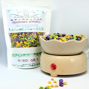 May include: A small bag of Mardi Gras-themed fragrance sprinkles, with a label that reads "SanJer Scents Fragrance Sprinkle" and "Mardi Gras Net Wt. 2 oz / 57 g". The sprinkles are in a ceramic bowl on a small electric warmer with a red light.