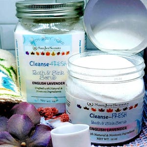 May include: Two glass jars of bath and sink scrub with a white powder inside. The jars have silver lids and labels that say "Cleanse-Fresh Bath & Sink Scrub" and "English Lavender". The labels also say "Crafted with Natural & Nontoxic Ingredients". The smaller jar is 12 oz and the larger jar is 24 oz.