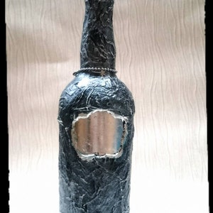 Op de afbeelding: Een grijze glazen fles met een getextureerd oppervlak en een zilveren label. De fles heeft een kristallen stop en een zilveren ketting om de hals.